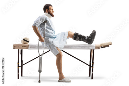 Bearded man in a hospital gown sitting at a medical table with a broken leg and neck collar