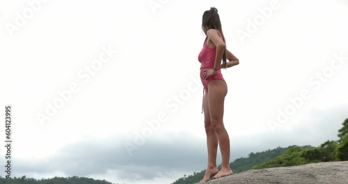 Attractive woman stands on rocks in bathsuit looking off into distance - wide shot photo