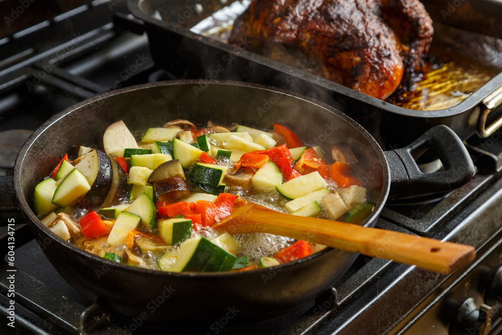 A frying pan with stewed vegetables on a plate next to a tray with baked chicken. A frying pan with stewed vegetables on a plate next to a tray with baked chicken.
