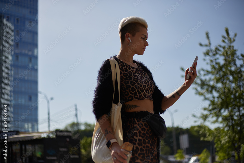 Stylish diverse female wearing leopard print clothes and tote bag on shoulder. Portrait of a short haired woman listening to music in wireless headphones and using a mobile phone outdoor