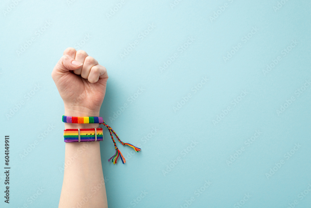 A raised hand wearing a rainbow-colored wristlets with a clenched fist symbolizing strength and the fight for rights takes center stage in this photo on pastel blue backdrop with space for text or ad - obrazy, fototapety, plakaty 