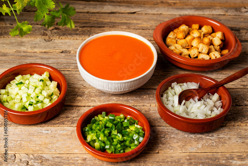 Bowl with gazpacho next to some clay plates with chopped vegetables and fried bread to add to the gazpacho, on a rustic table.