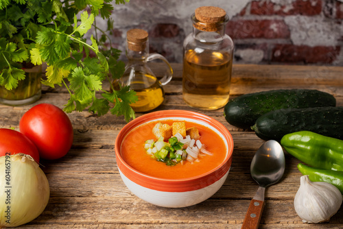 фотографія Vegetables necessary to make a gazpacho around a bowl full of gazpacho with chopped vegetables and chunks of toasted bread, on a rustic wooden table, diner view