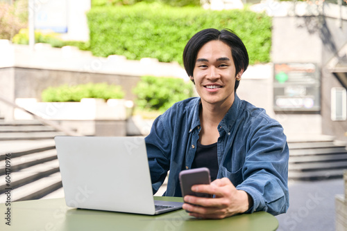 Young confident Japanese businessman using laptop holding mobile phone working online outdoors. Portrait of handsome successful asian freelancer sitting at workplace. Technology concept