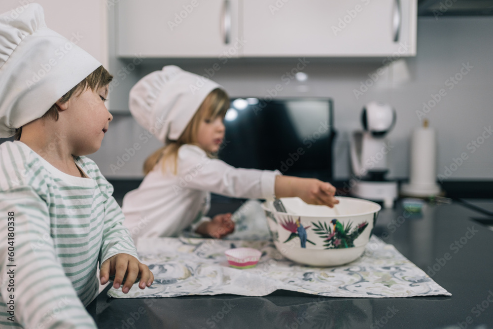Blond boy in a chef's hat watching his sister as she beats eggs in a bowl to cook homemade muffins in the kitchen. Blond boy in a chef's hat watching his sister as she beats eggs in a bowl to cook homemade muffins in the kitchen.