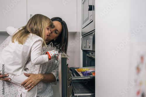 Mother holding her blonde daughter in her arms to peek at the tray of homemade cupcakes through the open oven door.