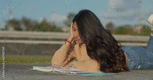 Young teenage Latina college student studying for an exam in an outdoor park with books