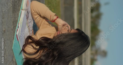 Young teenage Latina college student studying for an exam in an outdoor park with books, vertical shot
