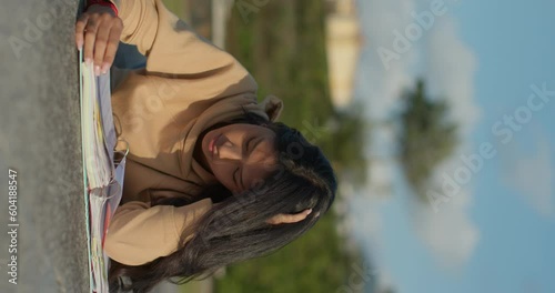 Young teenage Latina college student studying for an exam in an outdoor park with books, vertical shot