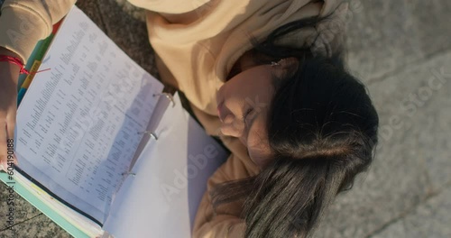 Young teenage Latina college student studying for an exam in an outdoor park with books, vertical shot