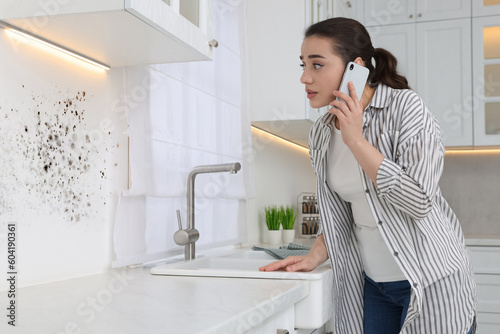 Mold removal service. Woman talking on phone and looking at affected wall in kitchen