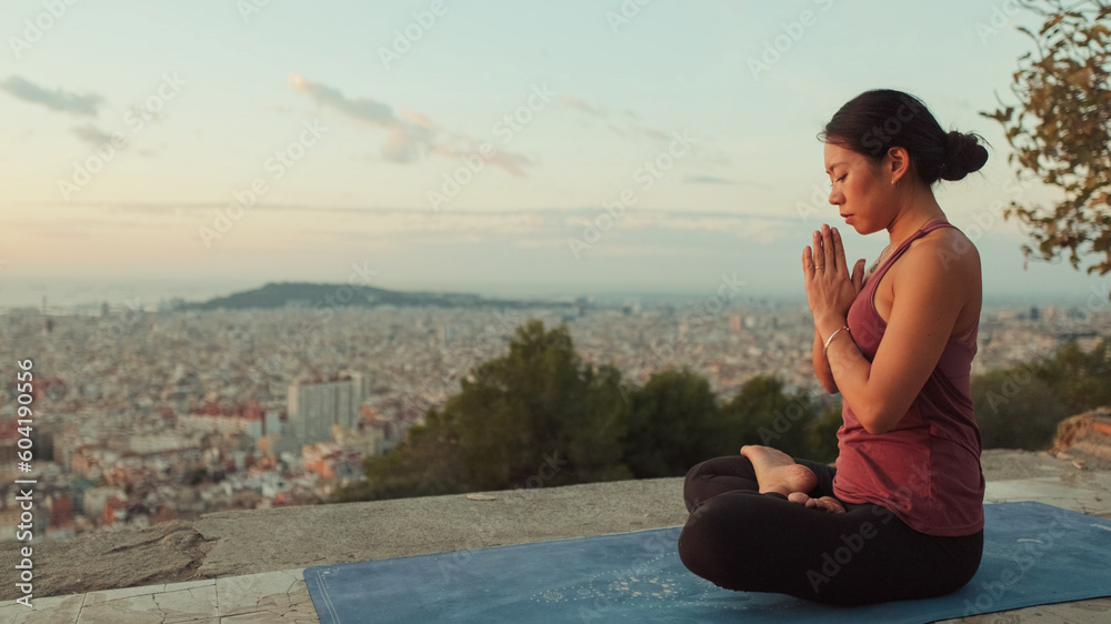 Young woman in bodysuit practices yoga at dawn at viewpoint. Girl raises her hands up while sitting in lotus position