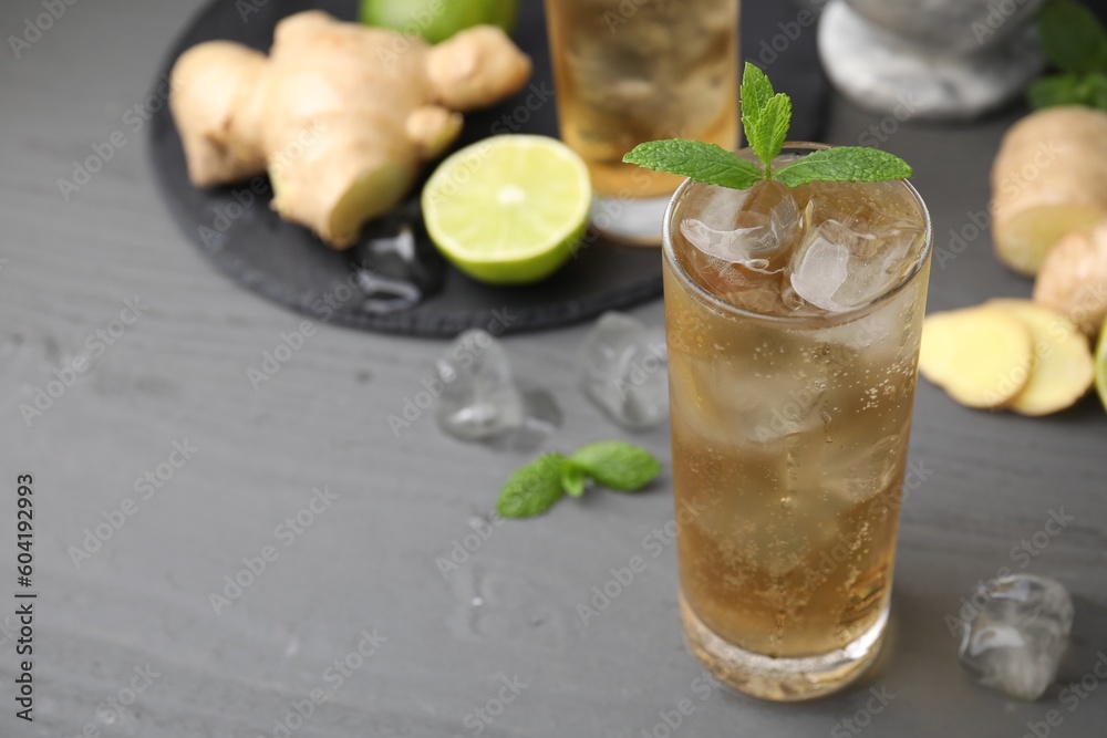 Glass of tasty ginger ale with ice cubes and ingredients on grey wooden table, closeup. Space for text