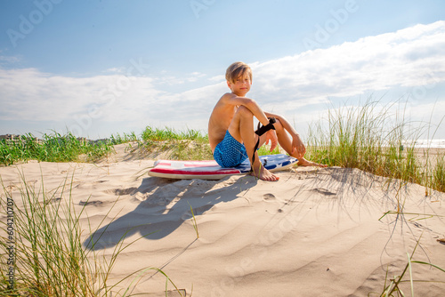 Foto Happy young boy sitting on sand dune at the beach leashed to his boogie board
