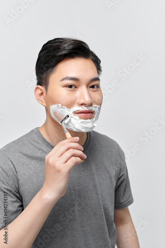Positive attractive young guy with gel foam on lower face shaving with razor at mirror