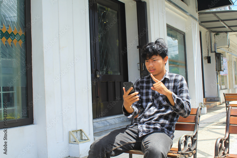angry young asian man pointing and threatening when have video call using his smartphone and sitting in the wooden bench