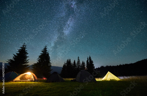 Night camping in mountains under starry sky. Tourist tents in campsite near burning campfire under beautiful sky full of stars with Milky way above forest. Concept of tourism and traveling.