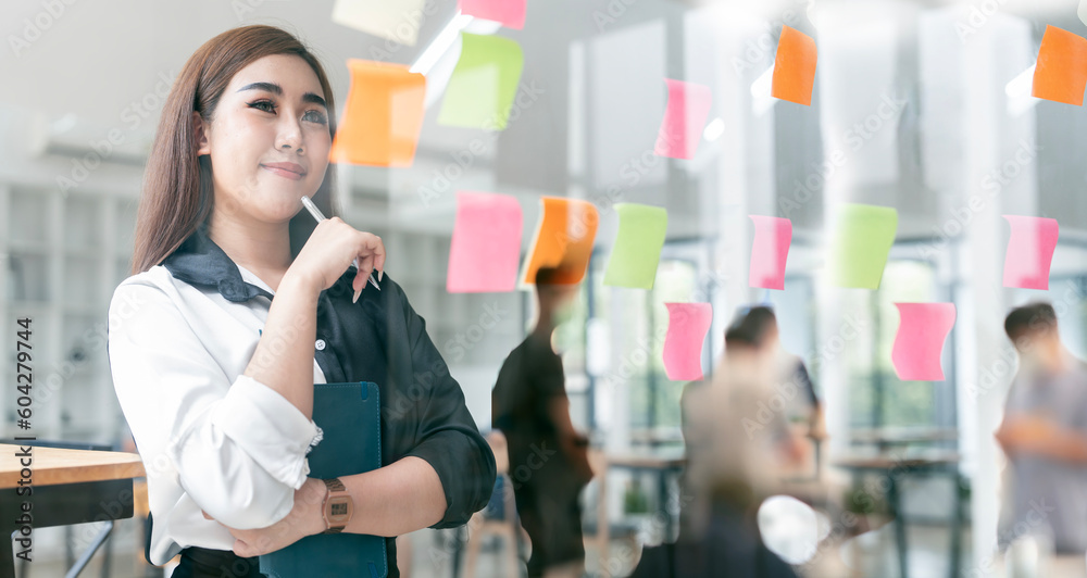 Smiling business people thinking and use sticky notes on glass wall in office with her colleague in background. Smiling business people thinking and use sticky notes on glass wall in office with her colleague in background.