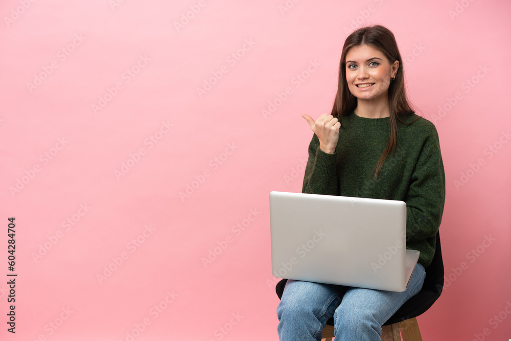 Naklejka premium Young caucasian woman sitting on a chair with her laptop isolated on pink background pointing to the side to present a product