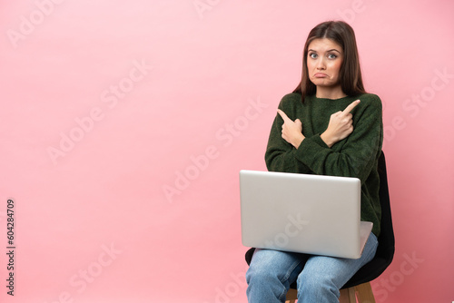 Young caucasian woman sitting on a chair with her laptop isolated on pink background pointing to the laterals having doubts