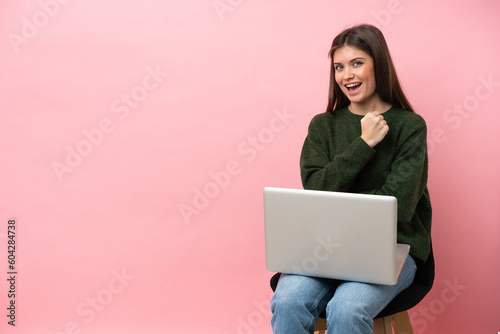 Young caucasian woman sitting on a chair with her laptop isolated on pink background celebrating a victory