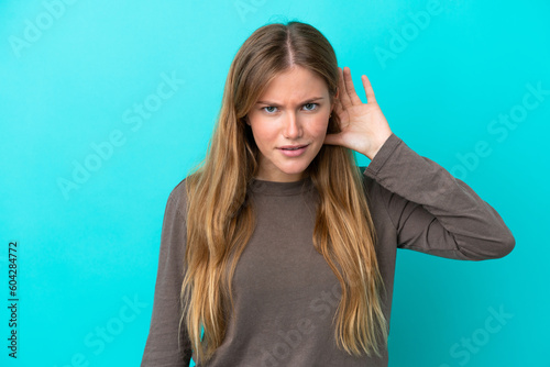 Young blonde woman isolated on blue background listening to something by putting hand on the ear