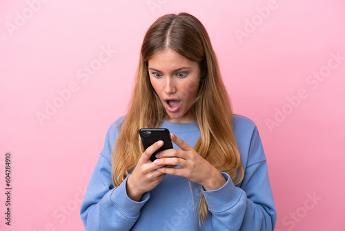 Young blonde woman isolated on pink background looking at the camera while using the mobile with surprised expression