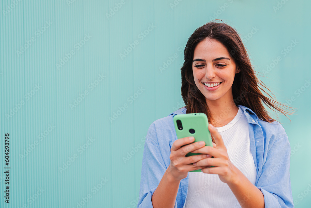 Smiling happy teenage woman using her smartphone and chatting online ,or browsing on internet at blue teal wall background. Young caucasian college student lady watching funny videos with mobile phone - obrazy, fototapety, plakaty 