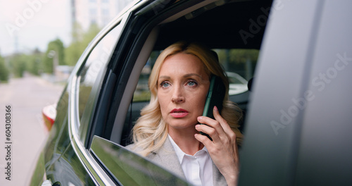 Successful business woman sitting in a luxury white car talking on the phone view from the street through the window. Lady manager in a suit. Communicates with partners. Business conversation. © serg