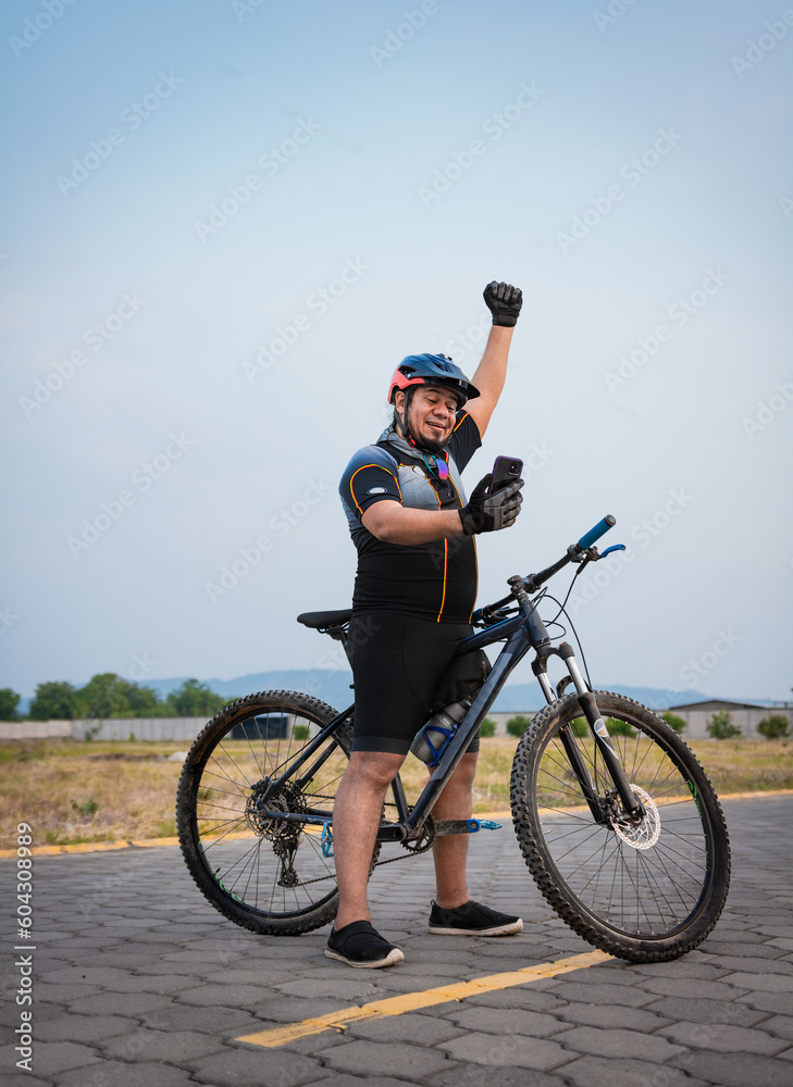 Fototapeta premium Professional cyclist looking happily at the cell phone in the street. Cyclist in sport suit using cell phone happily in the street