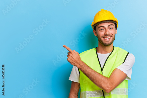 Cheerful bearded worker in yellow safety helmet and yellow vest smiles happily, points his finger out empty space for your ads, professional people concept, copy space
