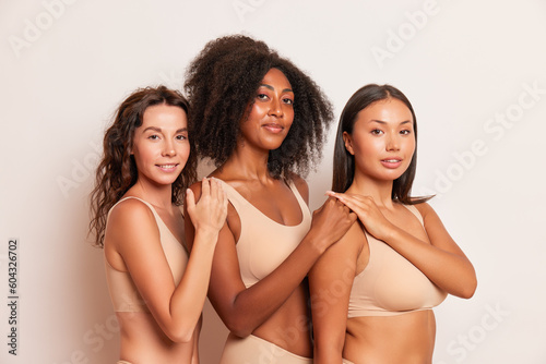 Three female models with different skin colours pose in the studio on white background, stand half-turned, each holds hand on the shoulder of friend, friendship concept, copy space © South House Studio