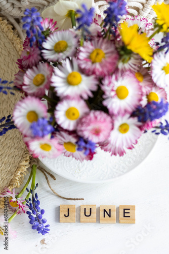 Bouquet of wild daisy flowers in a cup, straw hat, book. Cozy still life on the window of an old countryside house, summer cottage. Floral home decoration. Word June written in wooden letters