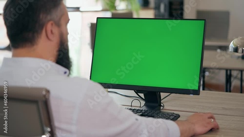 Over the Shoulder: Bearded Middle Aged Man Sitting at His Desk Uses Desktop Computer with Green Mock-up Screen. Male Manager or Businessman Works at Computer in Office