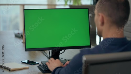 Over the Shoulder: Young Man Sitting at His Desk in Office Uses Desktop Computer with Green Mock-up Screen. Close up Green Screen Mock Up Display