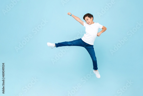 Cute mixed race Asian kid boy playing by jumping with wide open legs and raising fist in light blue isolated background studio shot