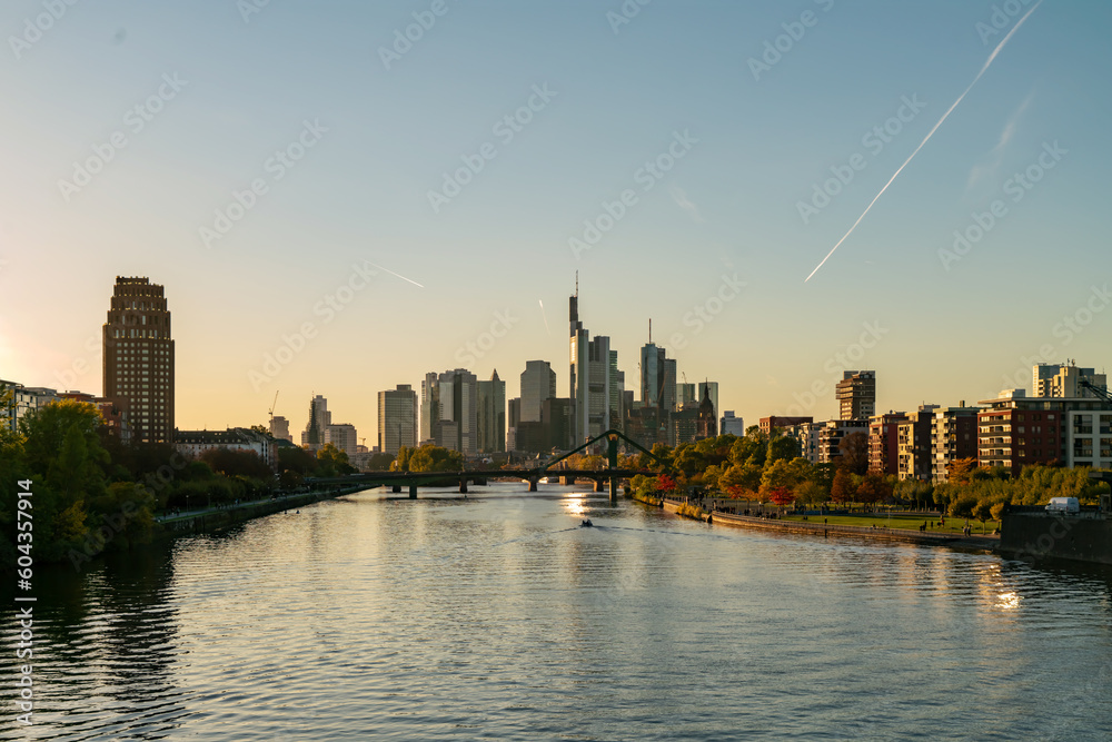 Obraz premium Stunning of Frankfurt's downtown skyline with a bridge, river reflections, and skyscrapers at sunset. Clear autumn sky and vibrant cityscape.