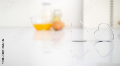 cooking and love concept - close up of home and heart shaped cookies cutter on white table with baking ingredients blurred background.