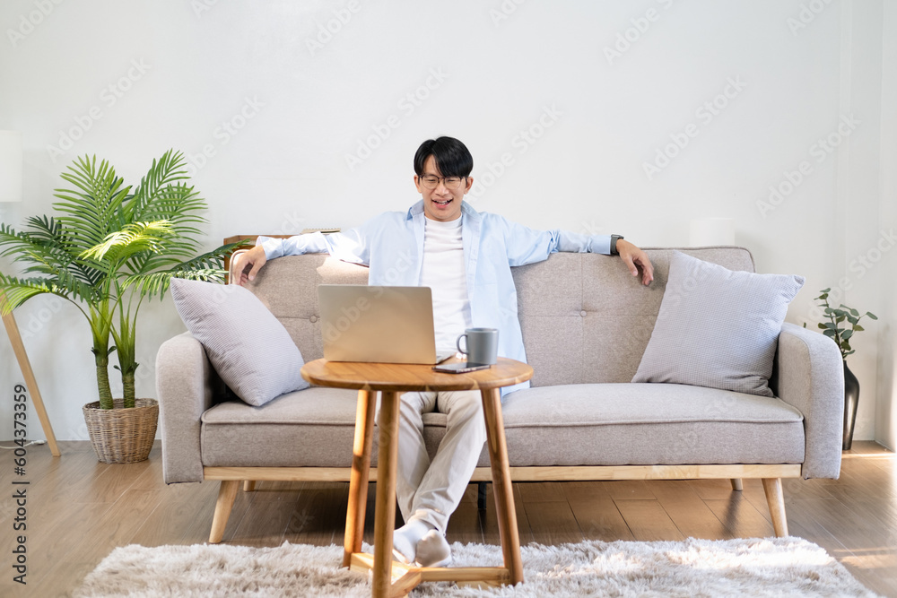 Smiling Asian businessman  sitting at office desk behind laptop. Happy employee feeling no stress, relaxing, watching funny video after successful working.