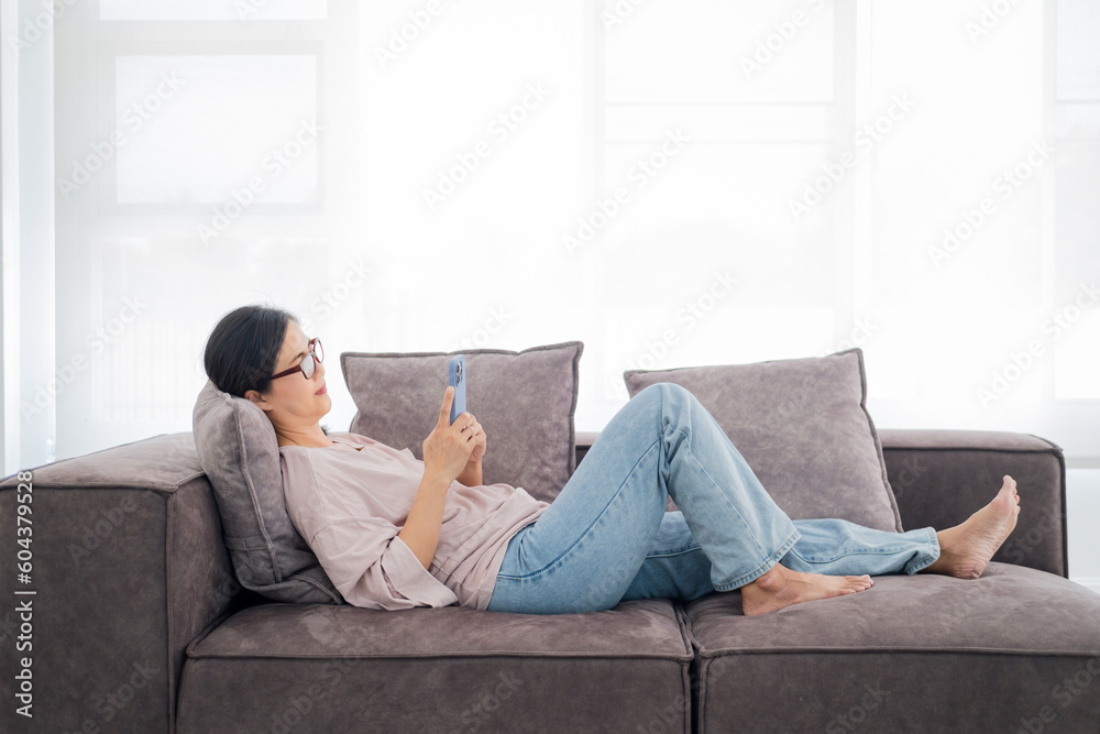 Happy mature asian woman relax on comfortable couch at home texting messaging on smartphone, smiling house wife use cell phone, browse wireless internet on gadget, shopping online from home Happy mature asian woman relax on comfortable couch at home texting messaging on smartphone, smiling house wife use cell phone, browse wireless internet on gadget, shopping online from home