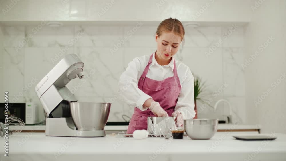 A female confectioner in a pink apron scoops chocolate dough with a plastic spatula from a food processor nozzle. Preparation for baking a dessert in a pastry shop.