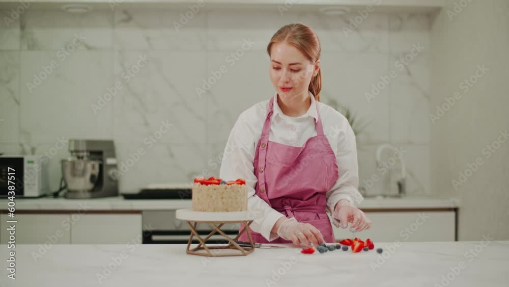 A female confectioner in a pink apron decorates the finished cheesecake with fresh fruits, strawberries and blueberries. A female confectioner in a pink apron decorates the finished cheesecake with fresh fruits, strawberries and blueberries.
