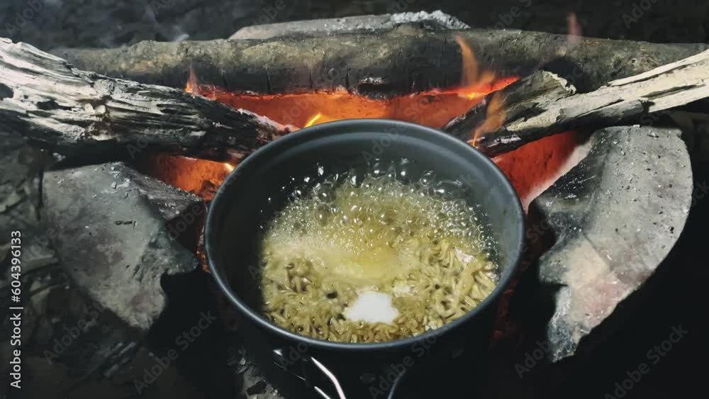 Boiling instant noodles and egg in a field pot on stove over a fire for cooking in a campsite. Bonfire cooking. Concept of travel and camping. Late night meal in camping. Boiling instant noodles and egg in a field pot on stove over a fire for cooking in a campsite. Bonfire cooking. Concept of travel and camping. Late night meal in camping.