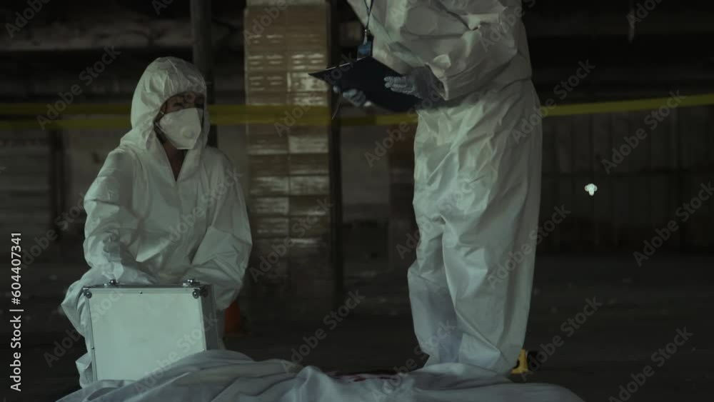 Full shot of forensic specialists in protective suits and masks working at murder scene, lifting blood-stained white sheet off dead body, discussing cause of death and writing on clipboard Full shot of forensic specialists in protective suits and masks working at murder scene, lifting blood-stained white sheet off dead body, discussing cause of death and writing on clipboard