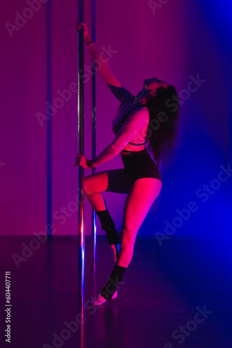 A girl dancer in a beautiful pose gracefully holds on to a pole in a gym with neon light. Sports pole dancing.