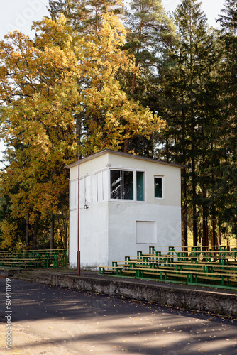 Rows of chairs and a commentator's booth of an abandoned stadium against the backdrop of autumn trees.