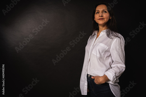 portrait of young girl about 40 years old, cute, emotional pensive, standing on dark background, white shirt and blue jeans, hand in pocket, studio shot with copy space