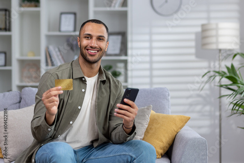 Online shopping. Portrait of a young African American man is sitting on the couch at home, holding a credit card and a phone. Smiling at the camera