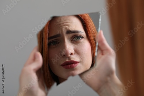 Young woman looking at herself in shard of broken mirror on light grey background, closeup photo