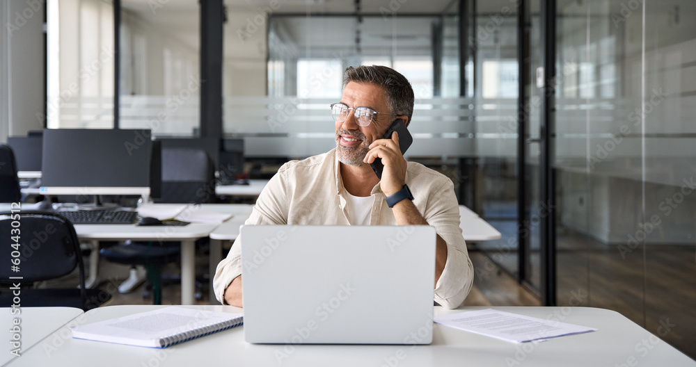 Middle aged Latin or Indian businessman having call on smartphone with business partners or clients. Smiling mature Hispanic man sitting at table talking by mobile cellphone at workplace in office. Middle aged Latin or Indian businessman having call on smartphone with business partners or clients. Smiling mature Hispanic man sitting at table talking by mobile cellphone at workplace in office.